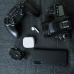 A collection of tech gadgets including a game controller, camera, smartphone, watch, and earphones on a fabric surface.